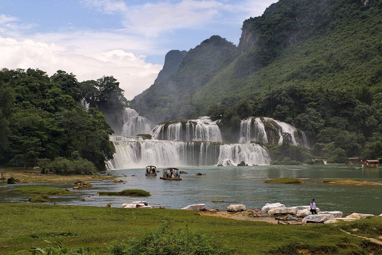 Ban Gioc waterfall pouring over wide limestone tiers into the Quay Son River, with bamboo rafts on the water and forested karst mountains under a partly cloudy sky