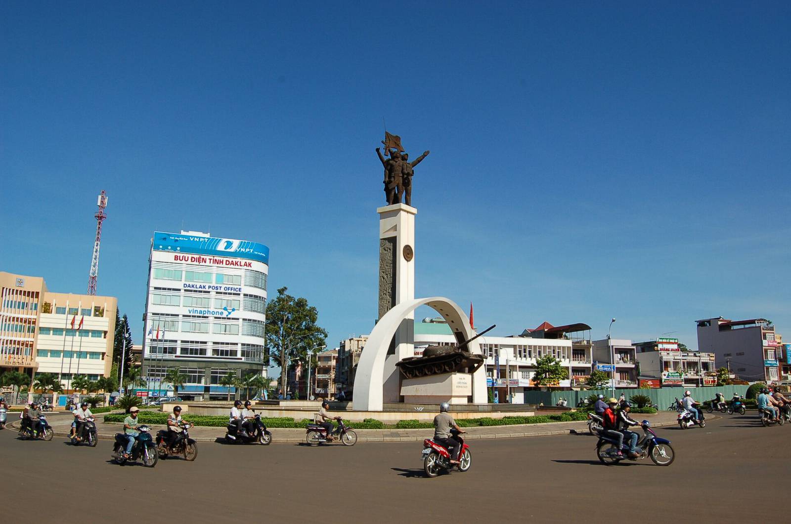 Buon Ma Thuot's Victory Monument, a bronze group of soldiers on a white column with an arch and a tank, at the central roundabout under a deep blue sky, with motorbikes circling
