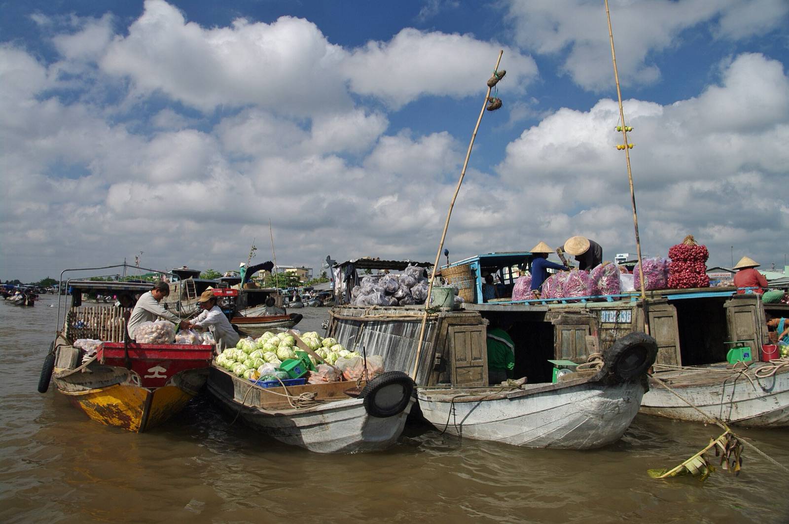 Wooden boats loaded with cabbages and onions at Cai Rang floating market near Can Tho, with tall poles showing goods for sale under a blue sky