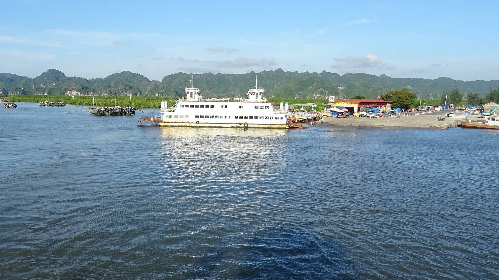 A white car ferry docked at the Cai Vieng pier on Cat Ba Island, with green limestone hills behind it, seen from a crossing ferry on a sunny afternoon