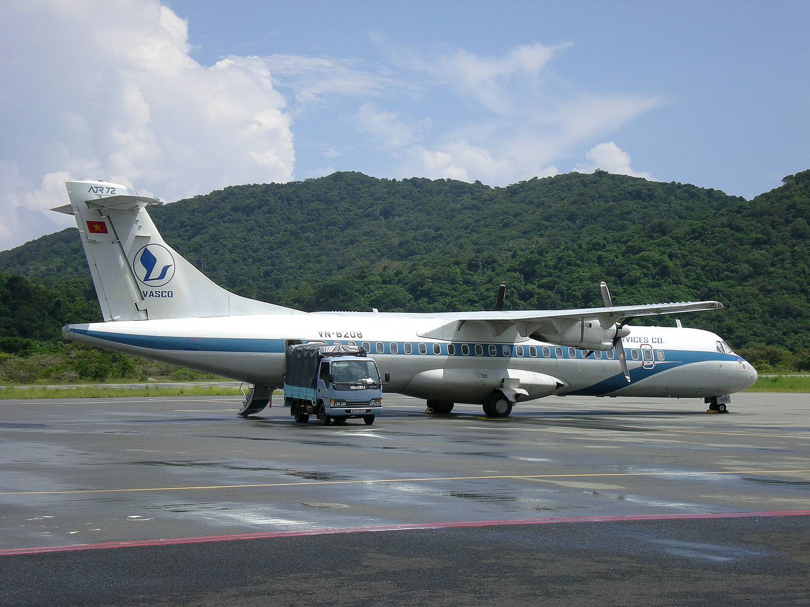 A VASCO ATR 72 turboprop parked on the apron at Con Dao airport, with forested green hills behind it under a partly cloudy sky
