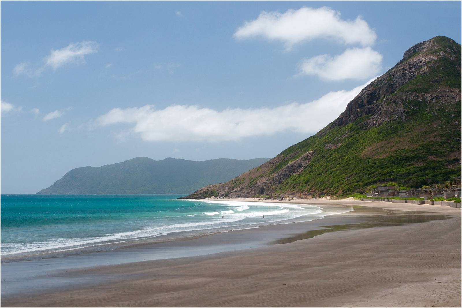 Dat Doc beach on Con Dao, a long curve of sand and turquoise water below a green mountain under a blue sky with white clouds