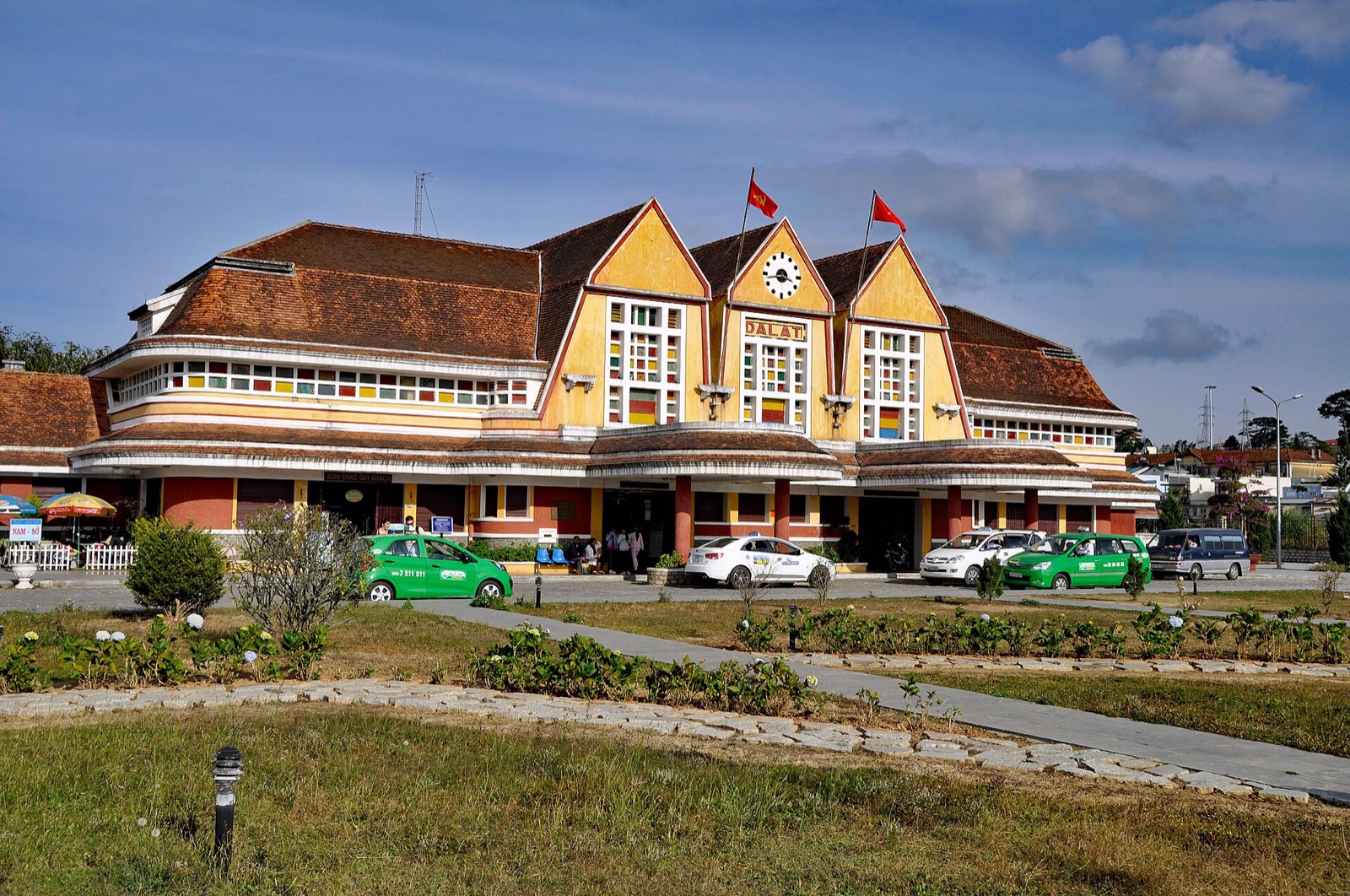 The yellow art deco Da Lat railway station with its three pointed gables and clock, seen across a lawn on a sunny day