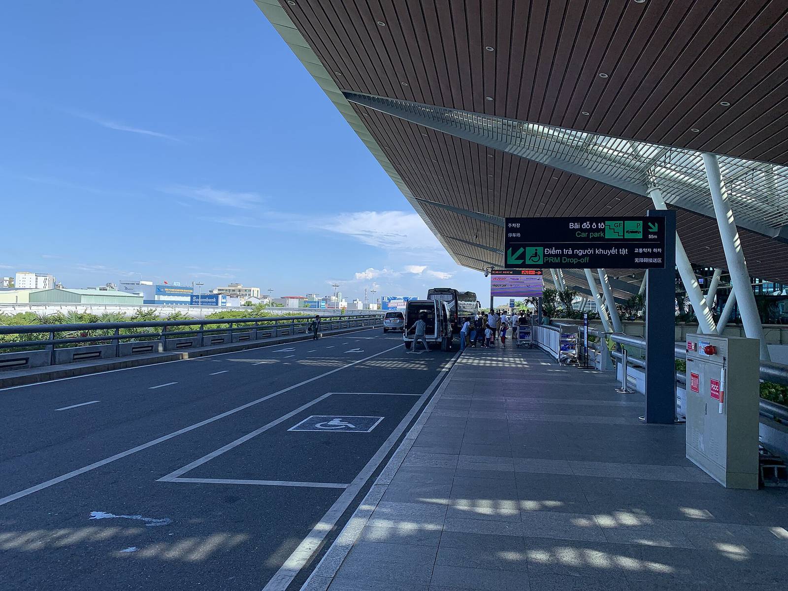 The departures kerb of Da Nang International Airport's international terminal under its long wooden canopy, with vans and travelers in bright afternoon sun