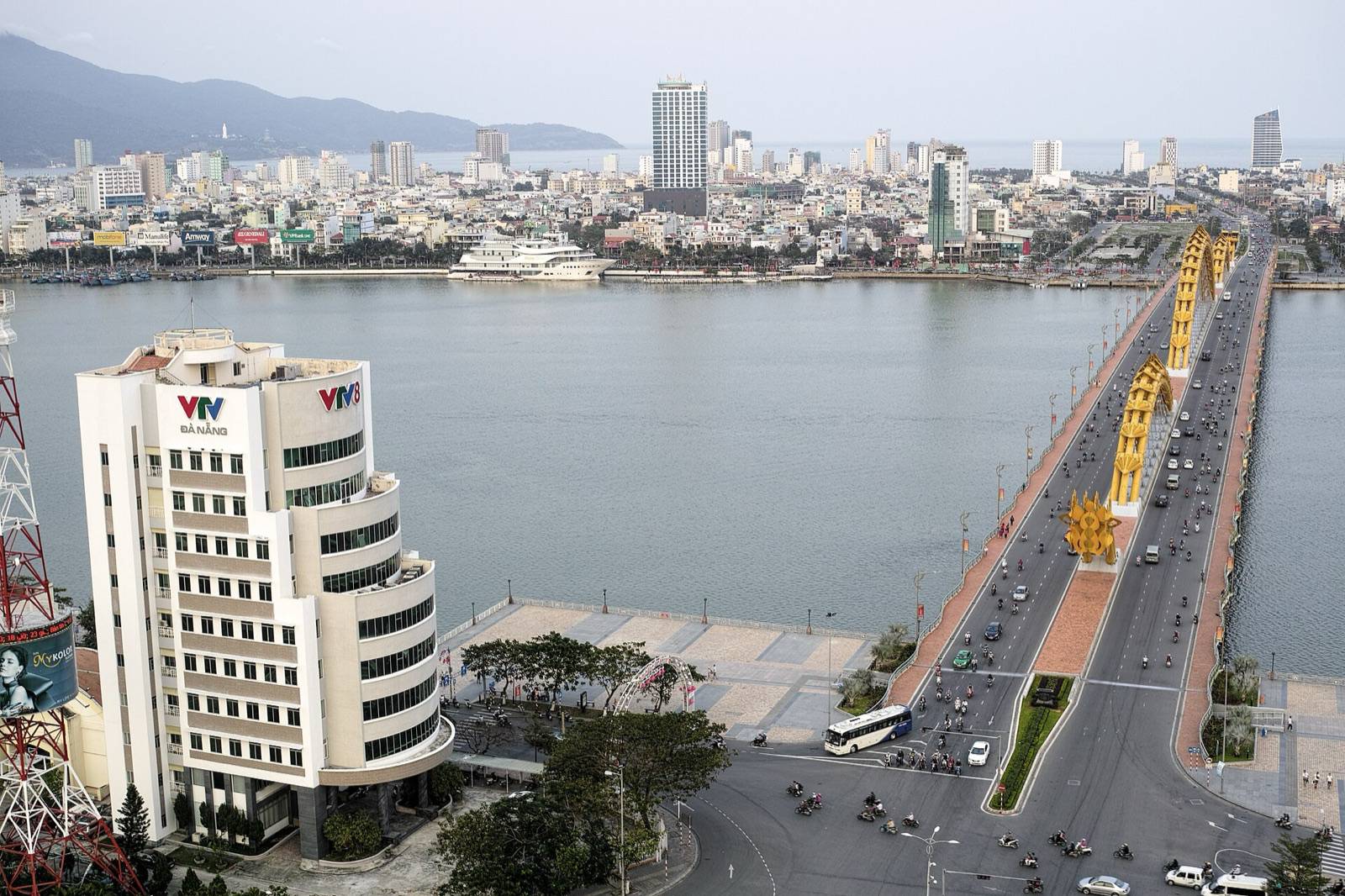 The yellow Dragon Bridge crossing the Han River in Da Nang on a bright day, seen from above the west bank with the city and coast beyond