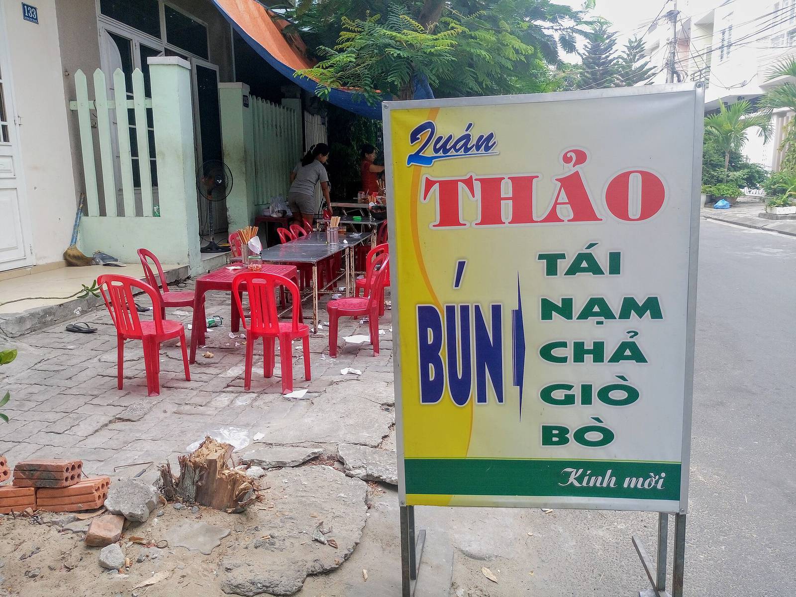 Red plastic chairs and steel tables at a sidewalk noodle eatery in Da Nang, beside a yellow menu sign for bun noodle soup, on a bright morning