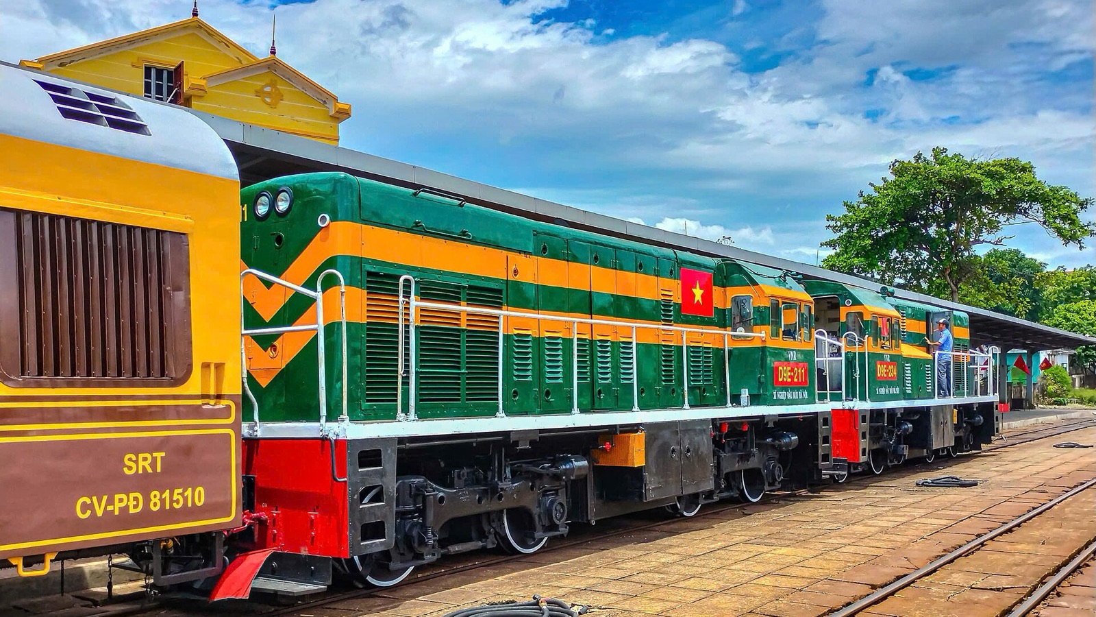 The green and orange locomotives of the Hue to Phong Nha heritage train standing at Dong Hoi station under a blue sky with white clouds