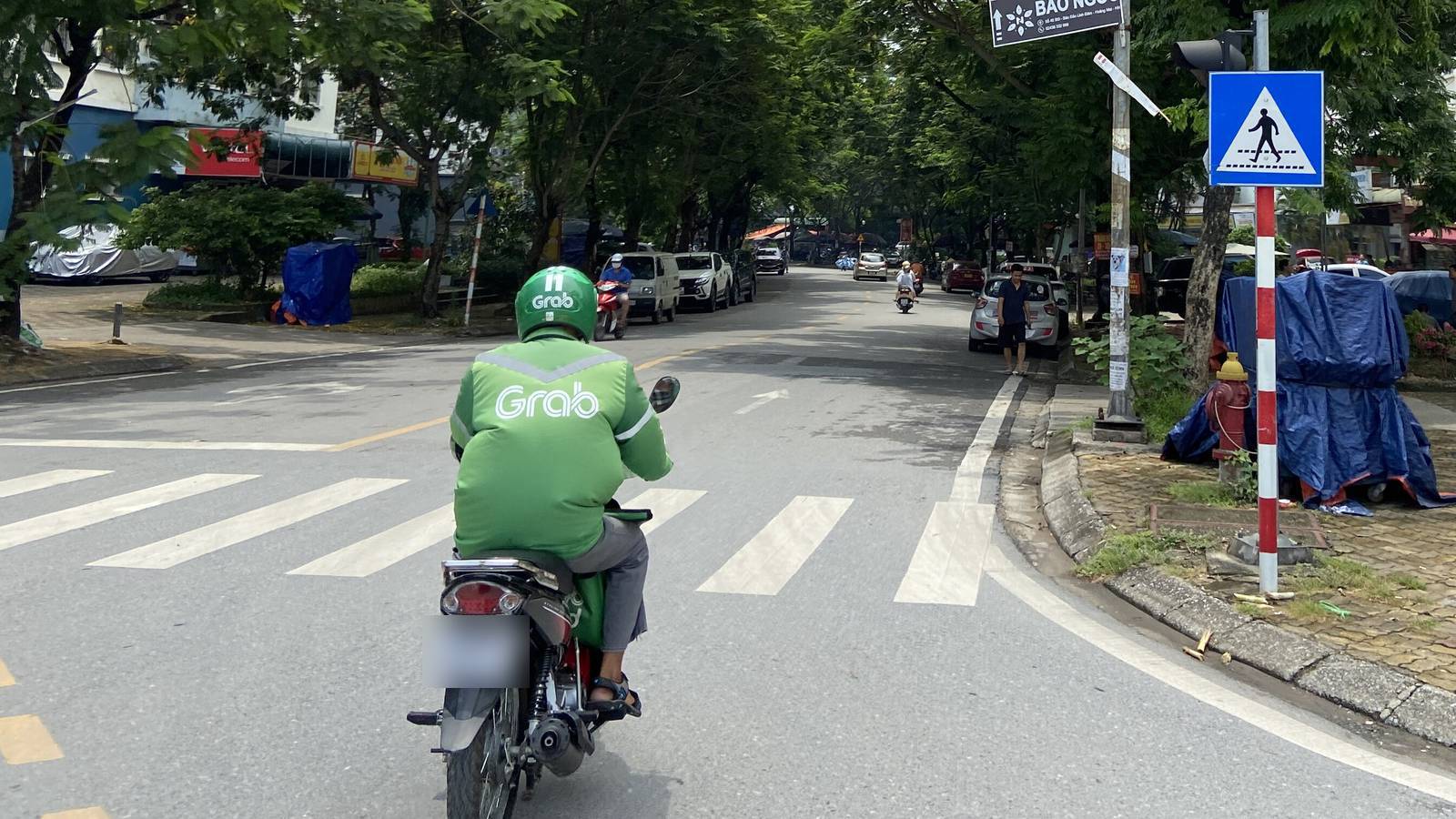 A Grab motorbike driver in a green jacket and helmet, seen from behind, rides across a zebra crossing on a leafy street in Hanoi on a bright day