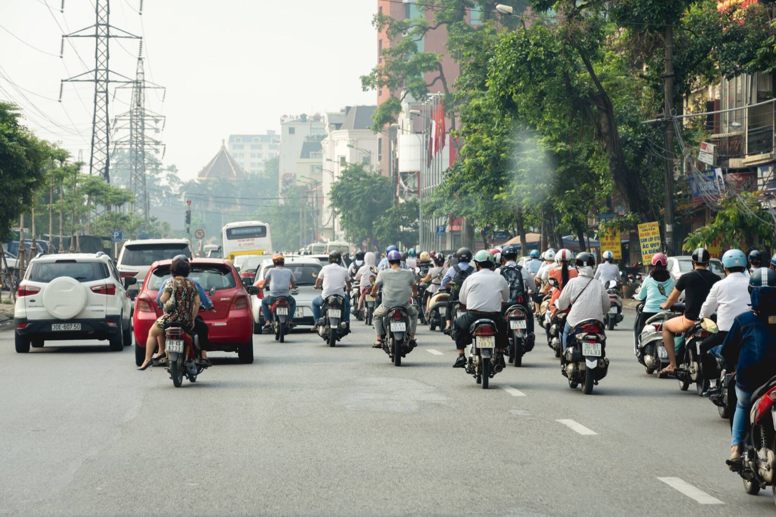 Dozens of motorbikes and a few cars head down a wide tree-lined avenue in Hanoi on a hazy morning, seen from behind