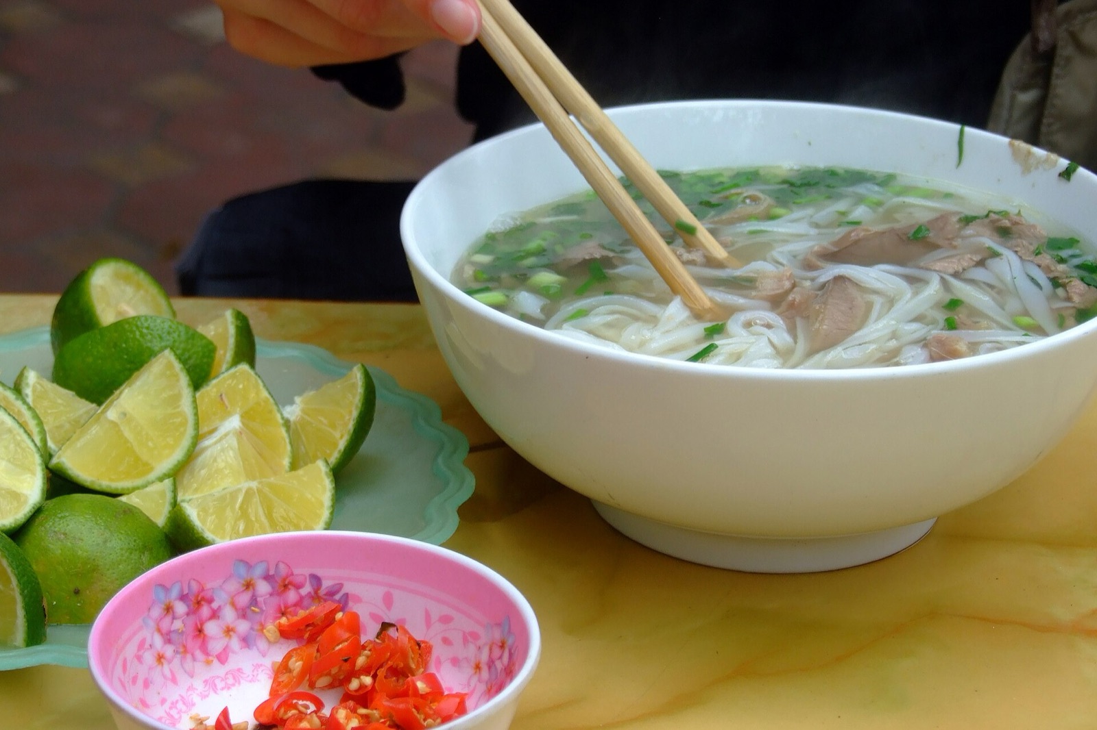 A steaming bowl of pho bo with rice noodles, sliced beef and spring onions beside a plate of lime wedges and a dish of fresh chili in Hanoi