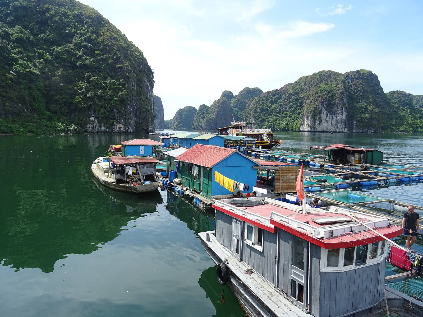 Blue and green floating houses and fish farms of a fishing village in Lan Ha Bay, with forested limestone islands under a bright sky