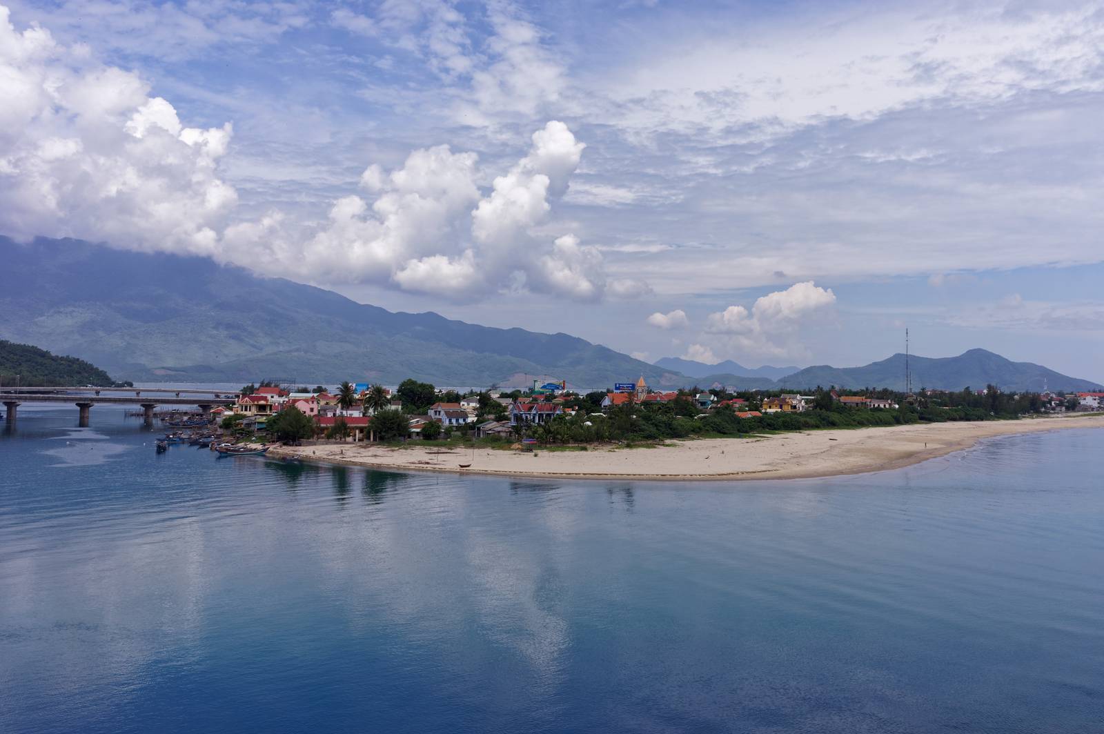 Lang Co village and its curving sandy shore beside calm blue water, with green mountains under white clouds on a bright day