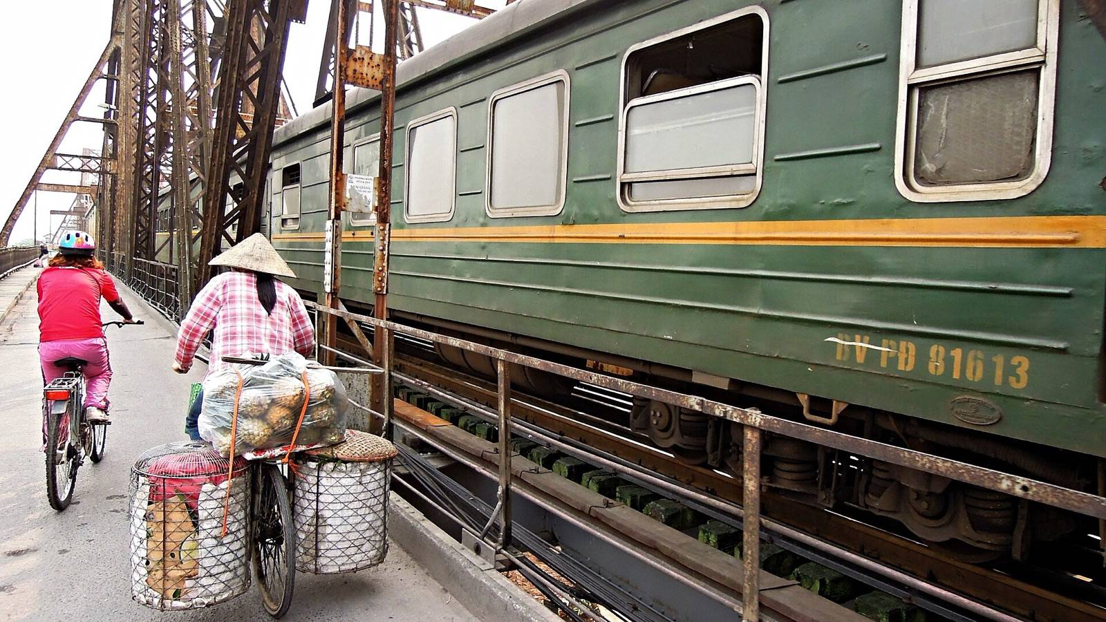 A green passenger train runs across Hanoi's rusting iron Long Bien Bridge while two cyclists ride along the bridge's side lane