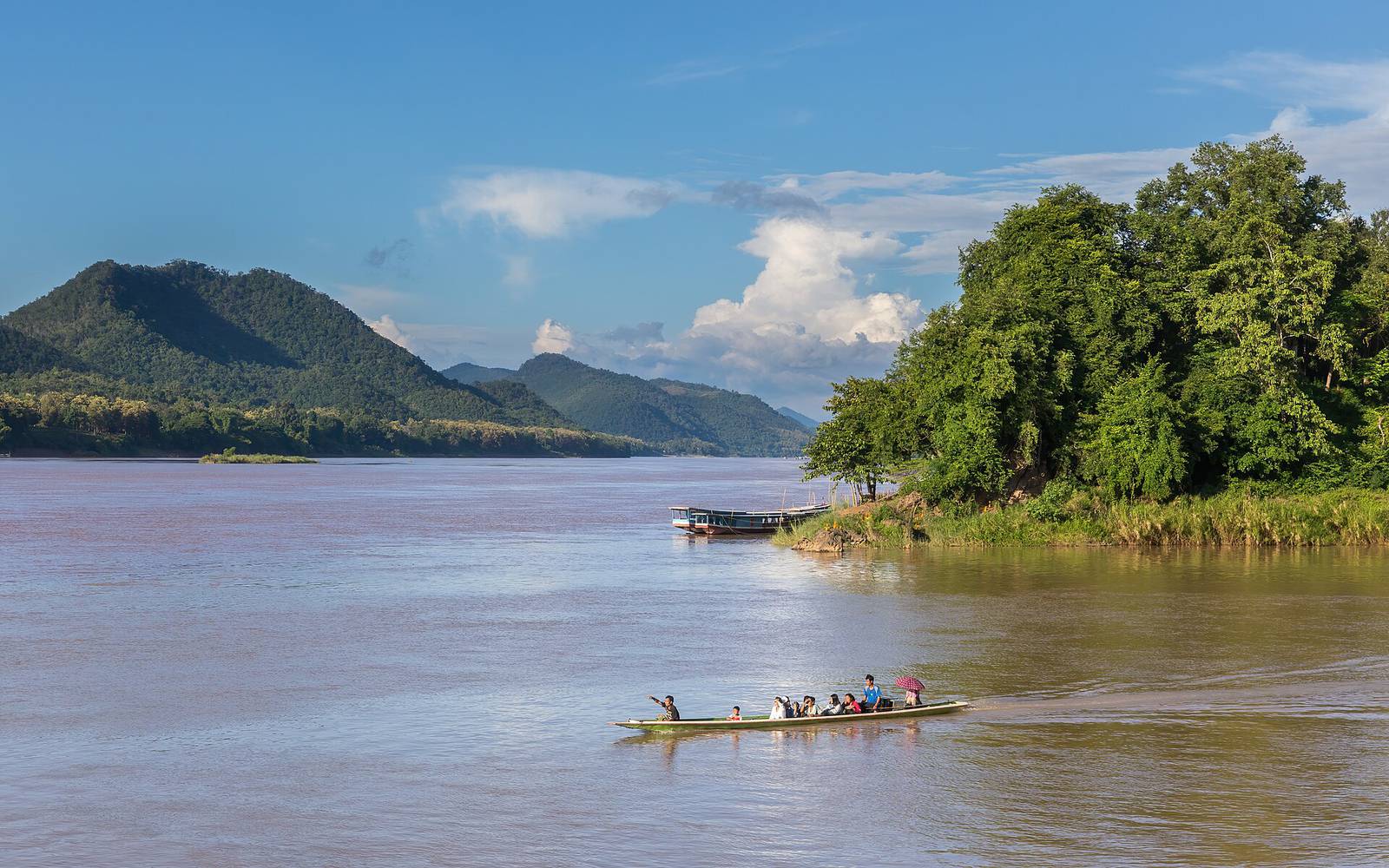 A long-tail boat with passengers crossing the brown Mekong where the Nam Khan joins it at Luang Prabang, with forested green hills under a blue sky with white clouds