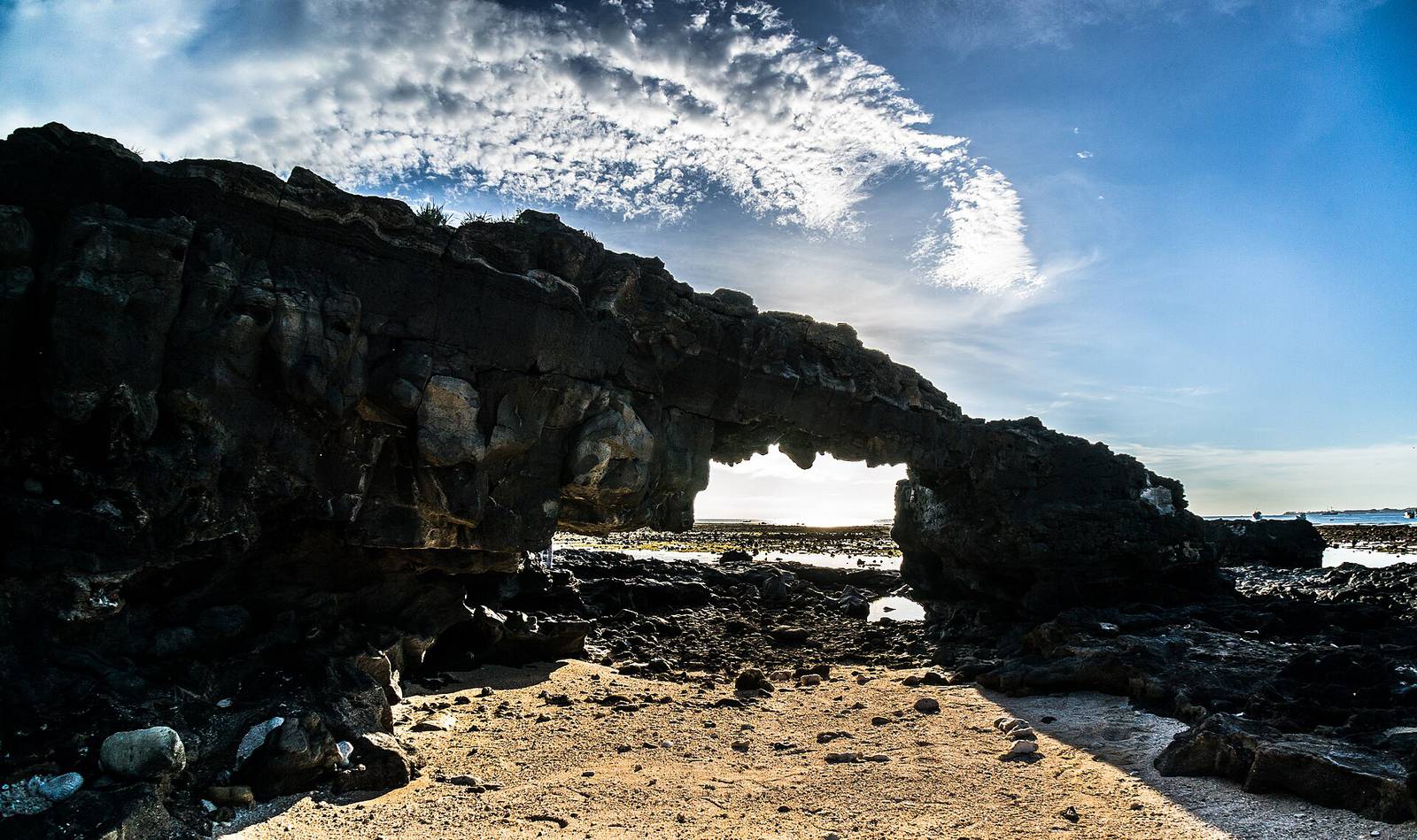 The To Vo rock arch on Ly Son island, a dark volcanic stone bridge over a rocky beach with blue sky and white clouds above