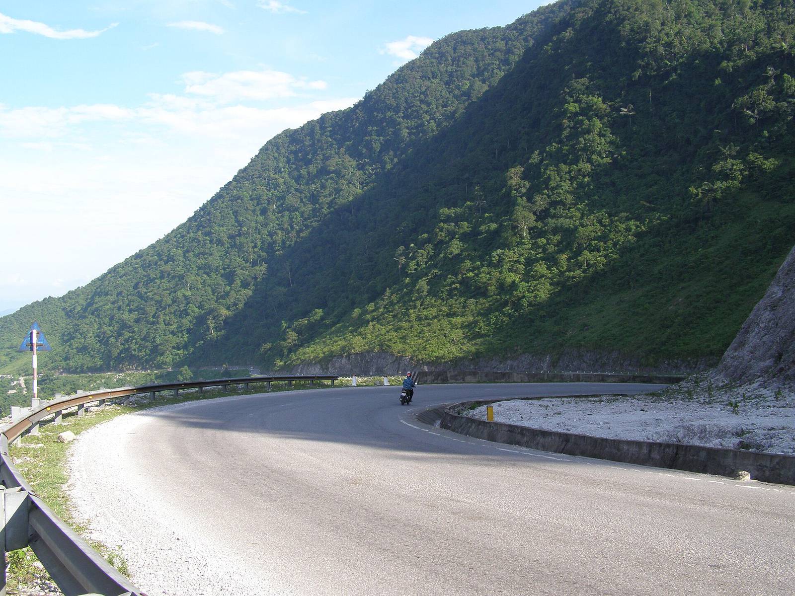 A sunlit hairpin bend edged with white limestone scree on the mountain road into Mai Chau, with a motorbike rounding the curve below a forested slope