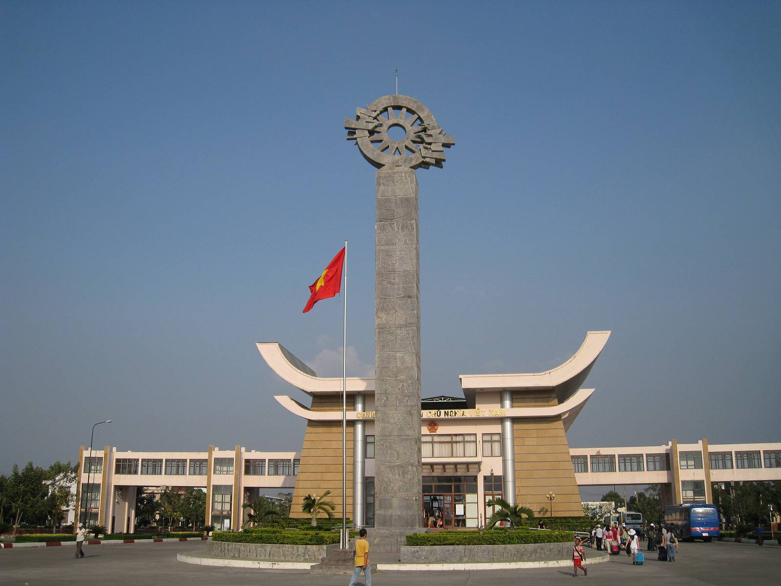 The Moc Bai border gate building in Tay Ninh with its curved roofs, a tall stone monument and a Vietnamese flag under a clear blue sky, with travelers and a bus in front