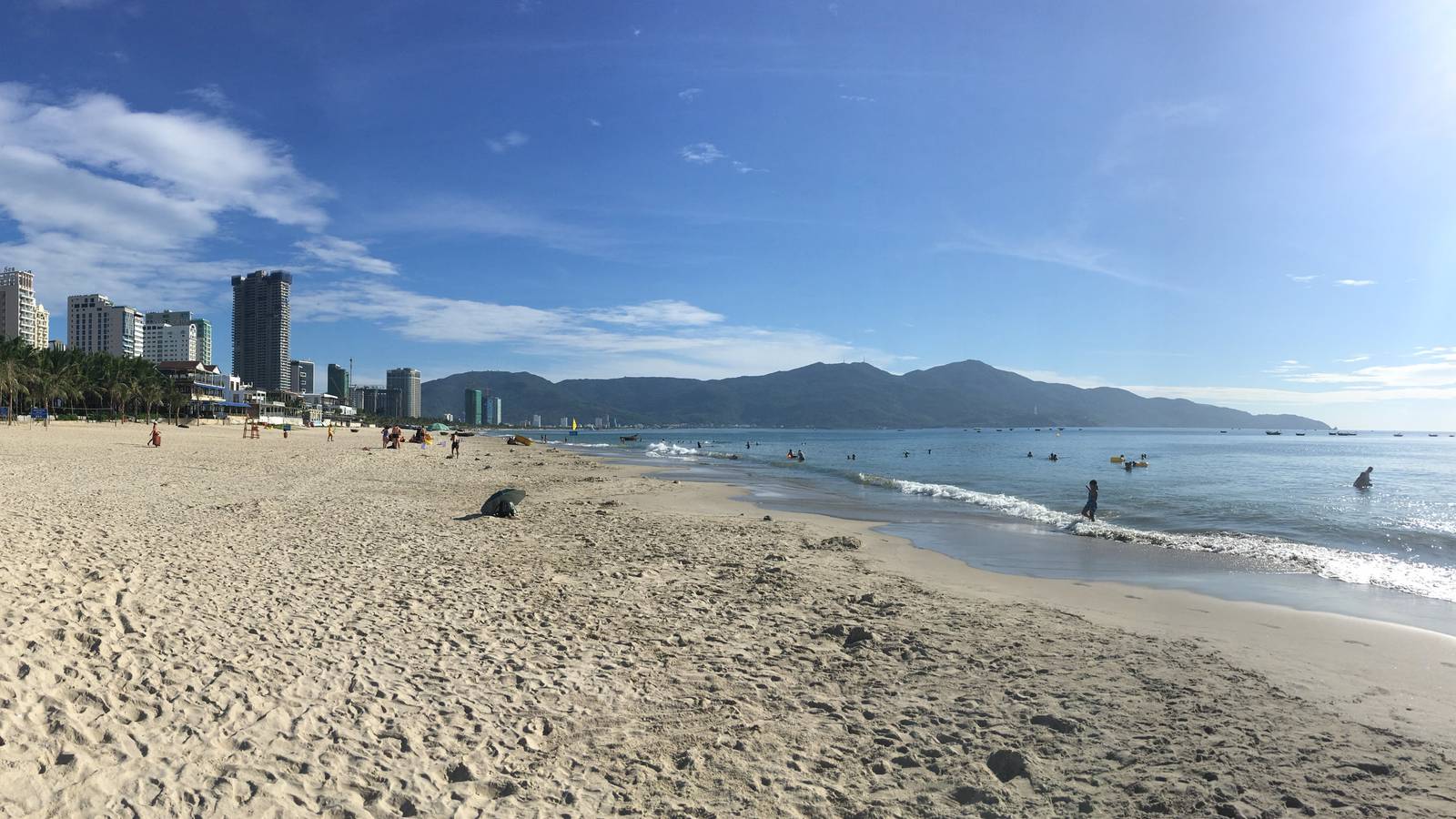Swimmers in the shallow surf and a few beachgoers on the wide sand of My Khe Beach in Da Nang, with Son Tra Mountain under a bright blue sky