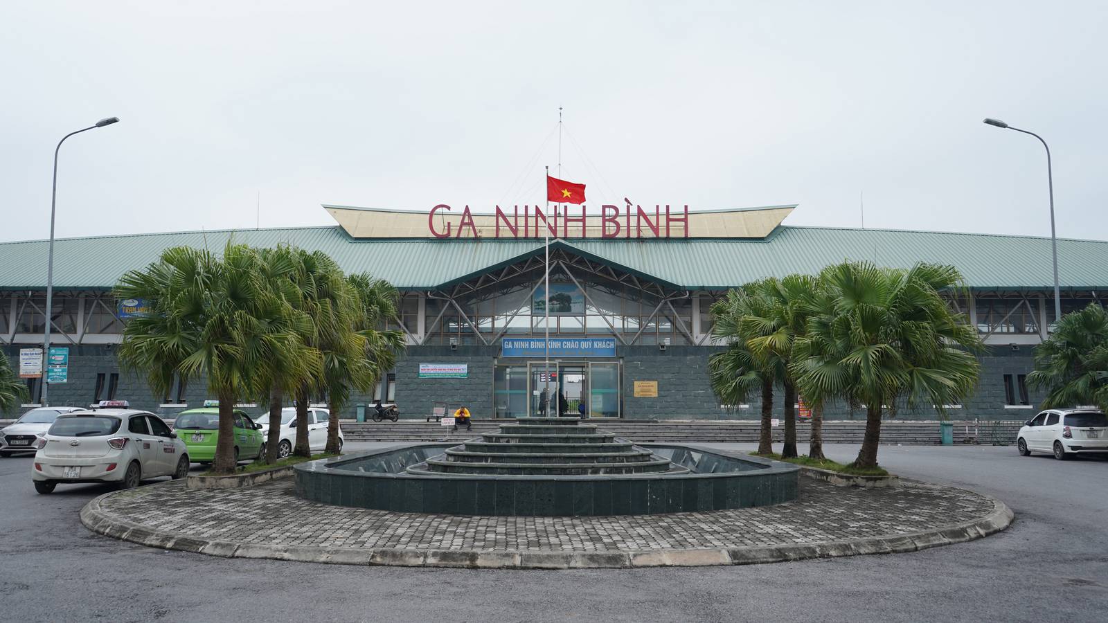 The front of Ninh Binh railway station, with its red GA NINH BINH sign, a Vietnamese flag, fan palms and waiting taxis on a pale, overcast day