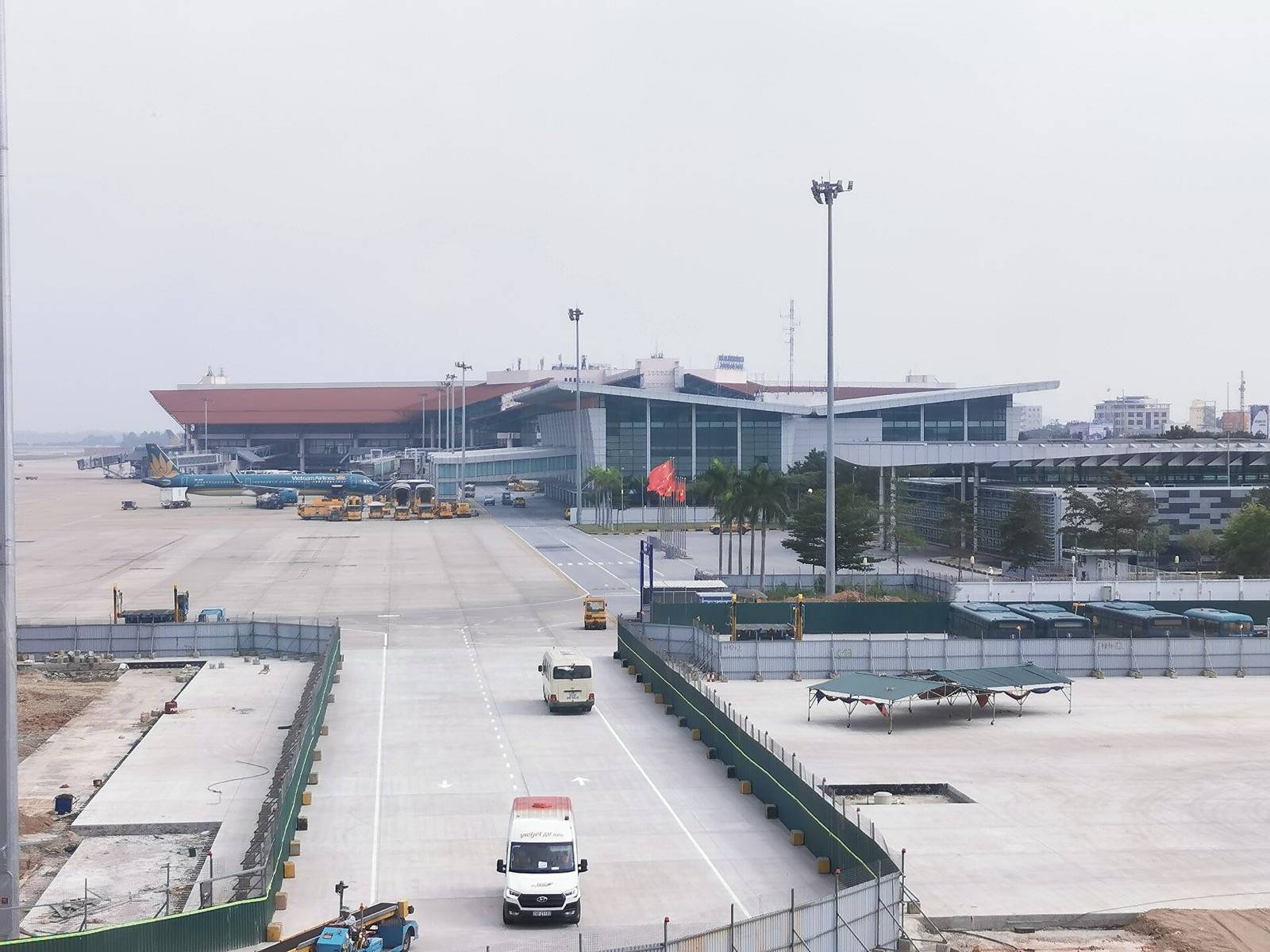 The terminal buildings of Noi Bai International Airport in Hanoi seen across the apron on a hazy day, with a parked airliner and service vans