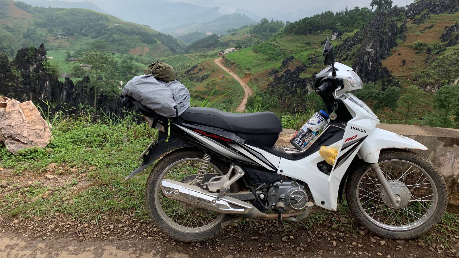A white Honda Blade semi-automatic motorbike with a bag strapped behind the seat, parked on a gravel roadside above a green karst valley in Vietnam