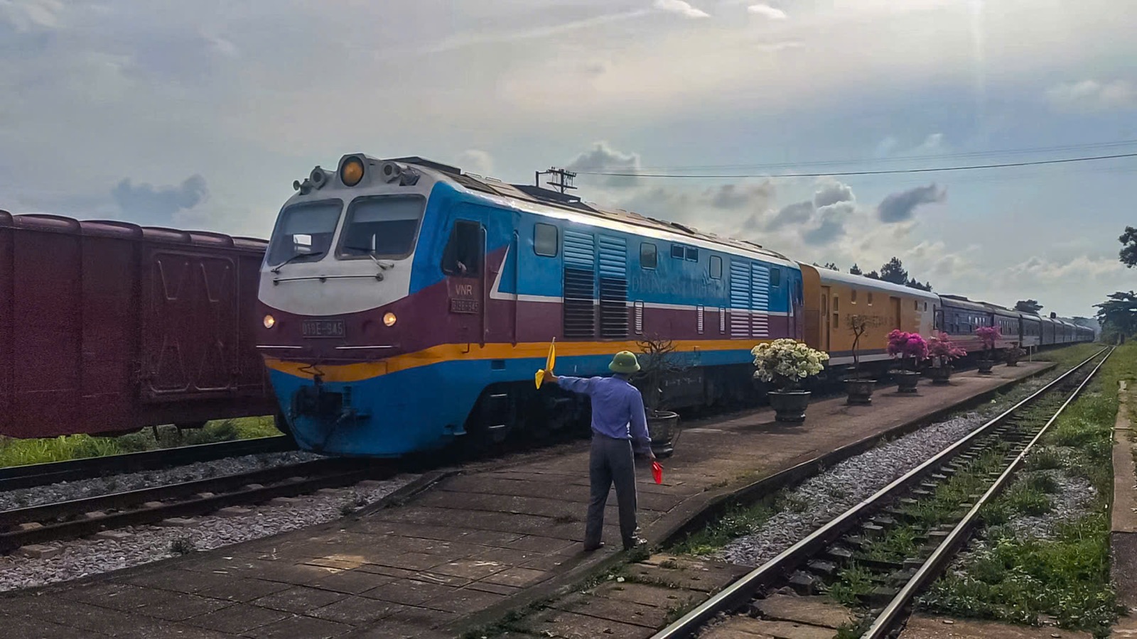 A blue and yellow diesel locomotive hauling the SE7 Hanoi to Saigon express through Tho Loc station on a bright afternoon, with a railway worker signalling from the platform