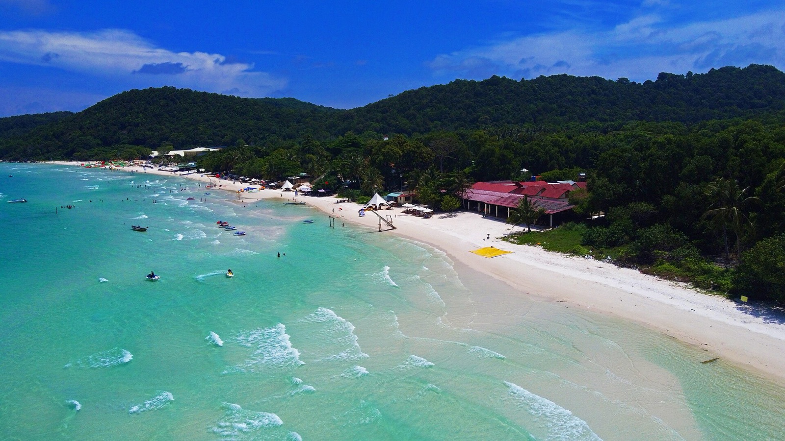 Aerial view of Sao Beach on Phu Quoc, with white sand curving along turquoise water below forested hills on a sunny day