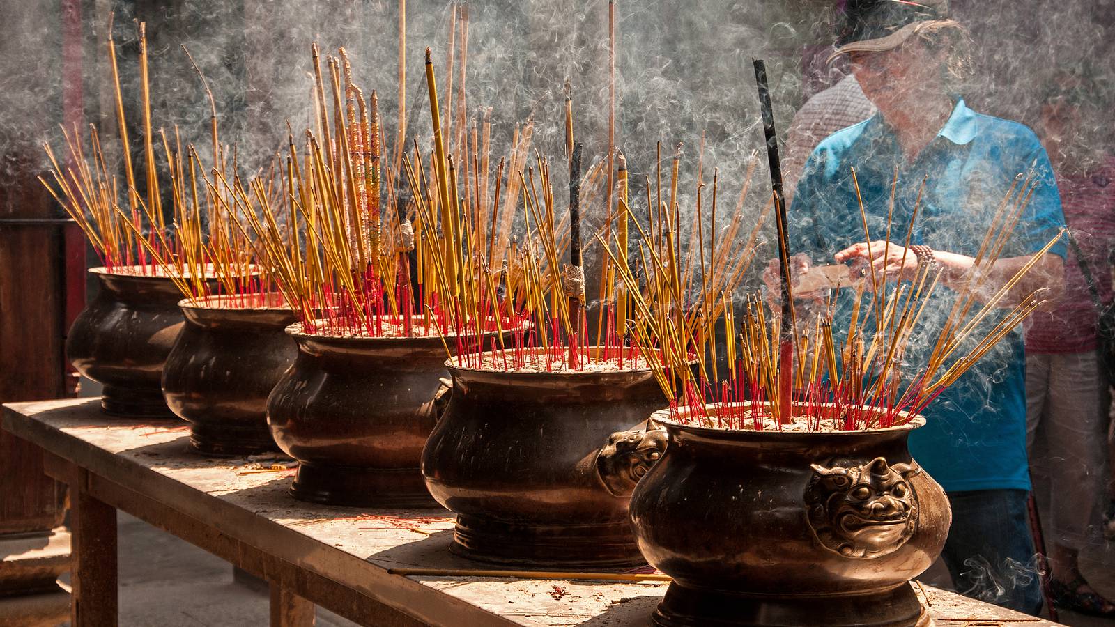 Incense sticks smolder in a row of bronze urns at Thien Hau Temple in Ho Chi Minh City while a worshipper in a cap adds more in bright sunlight