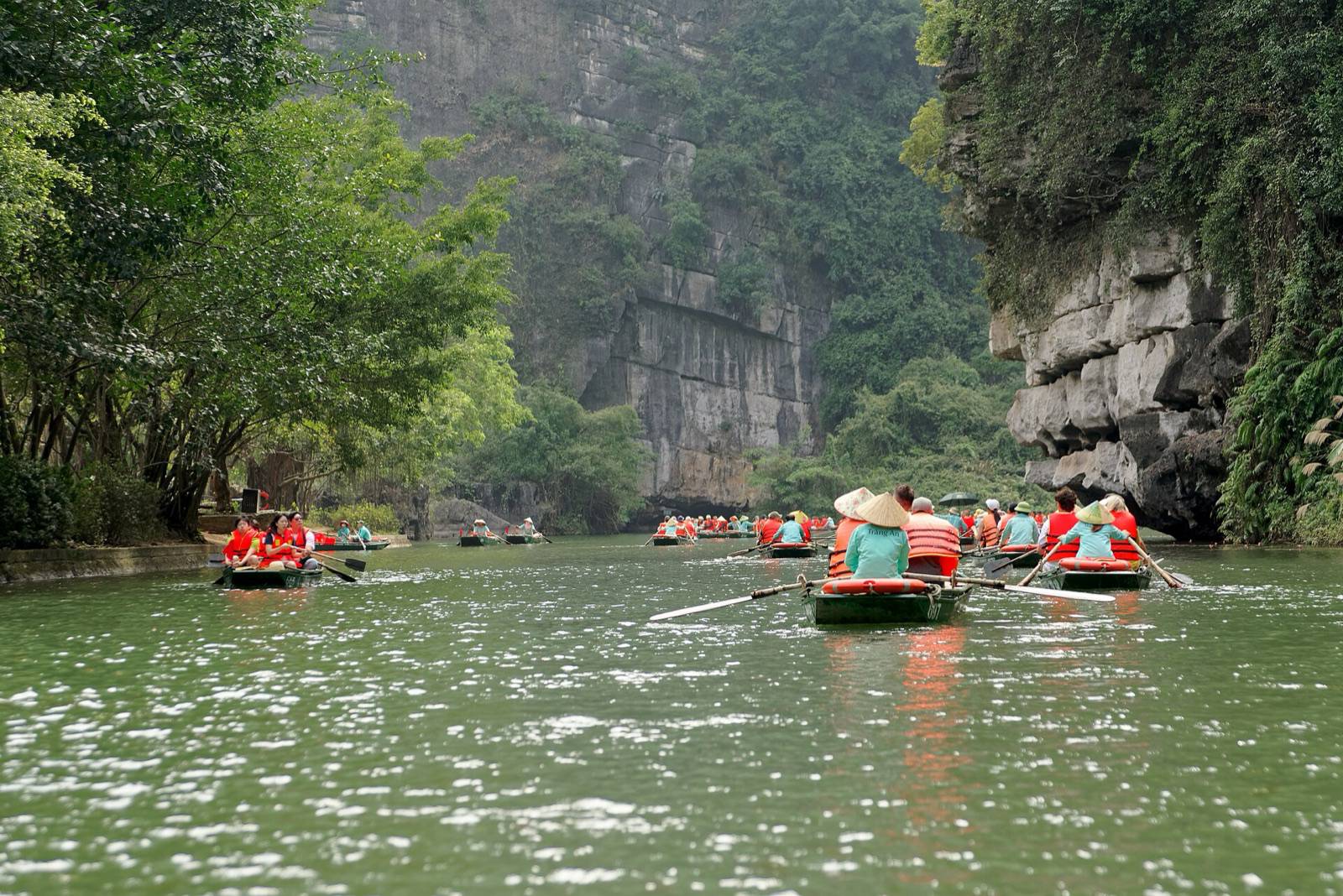 Rowing boats with tourists in orange life jackets on the river at Trang An, between limestone karst cliffs in Ninh Binh