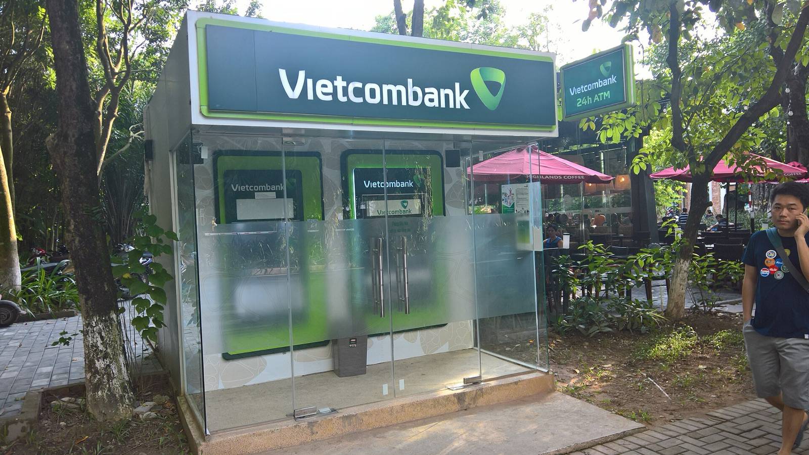 A glass 24-hour ATM kiosk with two cash machines under green bank signage beside a cafe terrace near Hanoi on a sunny day