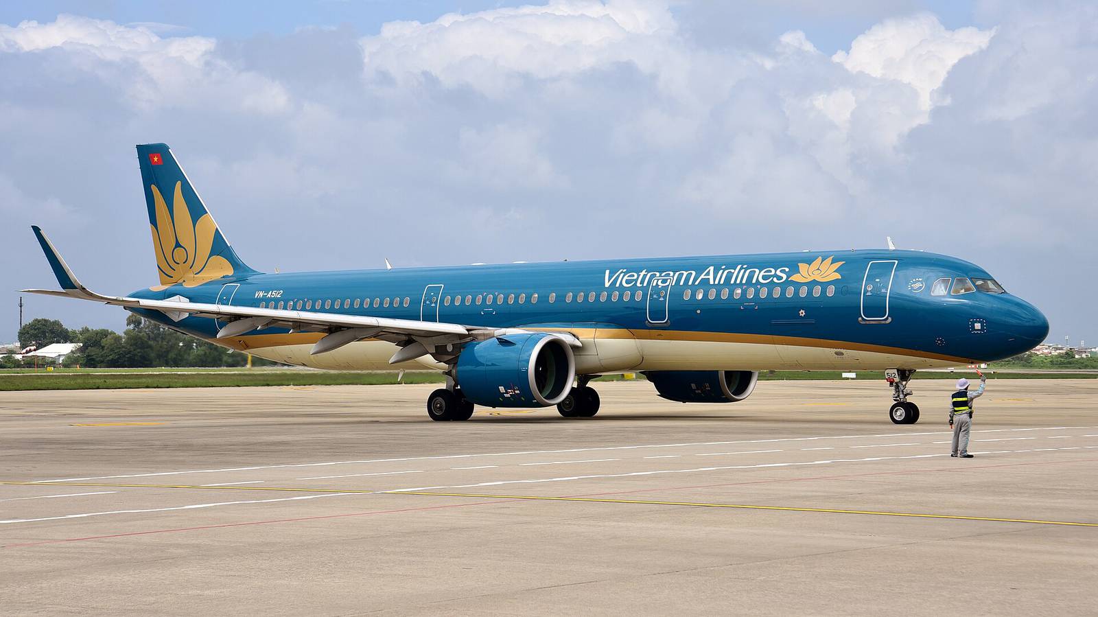 A blue Vietnam Airlines Airbus A321neo taxiing toward the domestic terminal at Da Nang International Airport under a bright, cloudy sky, guided by a ground marshal