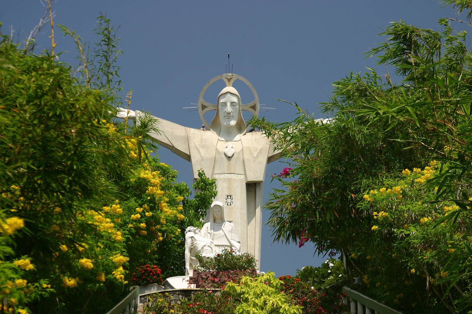 The white Christ the King statue on Nui Nho in Vung Tau, arms outstretched above flowering trees and a Pieta sculpture, under a deep blue sky